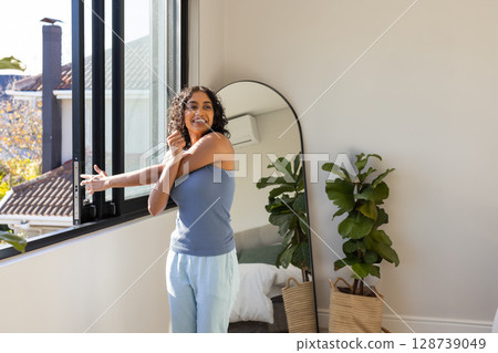Indian woman stretching arm while opening sliding window in bright bedroom, with mirror and plants 128739049