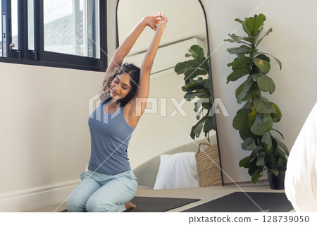 Indian woman in 20s kneeling stretching overhead on yoga mat near window in bedroom, copy space 128739050