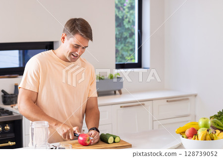 Young adult male slicing apple on cutting board beside blender jar at kitchen island, copy space 128739052