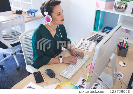 Woman typing on keyboard at desk with monitor in modern office, wearing headphones Woman typing on keyboard at desk with monitor in modern office, wearing headphones 128739068