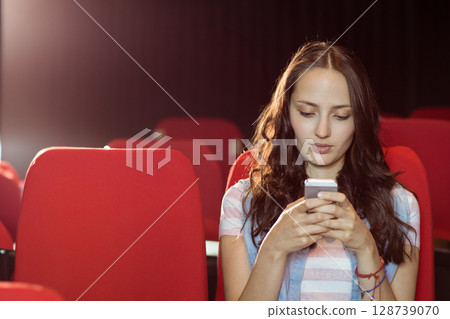 bi-racial young adult woman sitting alone in red auditorium seats using smartphone, copy space 128739070