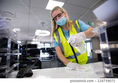 Woman wearing safety vest cleaning office desk with disinfectant bottle and cloth in modern office 128739072