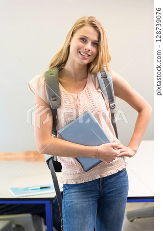 woman standing in classroom area, holding closed laptop and gray backpack, displaying notebook pen 128739076