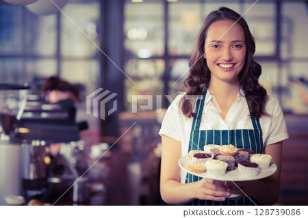 Young female barista holding plate of mixed cupcakes in cafe with espresso machine, copy space 128739086
