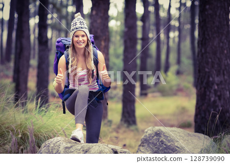 Woman standing on large rock in pine forest, carrying purple backpack and wearing knit pom-pom hat 128739090