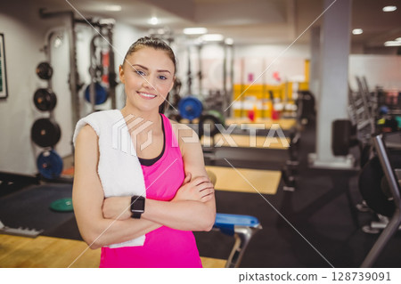 Young adult woman standing with arms crossed at gym, holding towel and wearing smartwatch 128739091