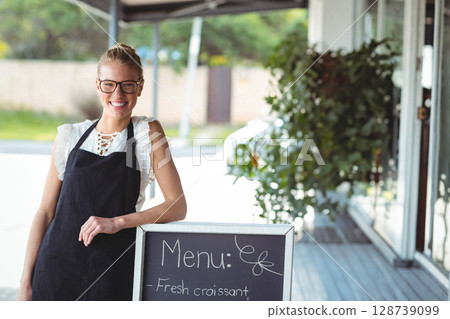 Woman smiling and leaning on chalkboard menu sign under cafe awning, wearing apron and glasses Woman smiling and leaning on chalkboard menu sign under cafe awning, wearing apron and glasses 128739099