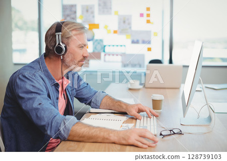 Middle-aged man typing on white keyboard in office by window, with headset, coffee cup, copy space 128739103