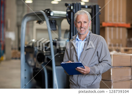 Senior man standing in warehouse holding clipboard and pen near forklift and pallets, copy space 128739104