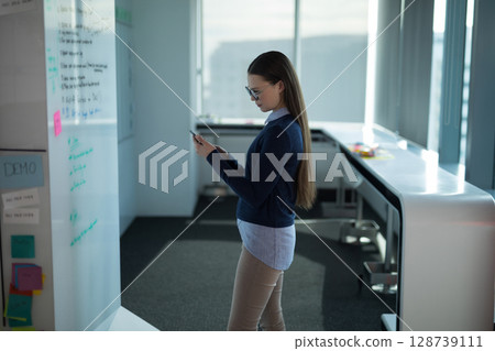 Young woman standing side-on checking smartphone in modern office near whiteboard with sticky notes Young woman standing side-on checking smartphone in modern office near whiteboard with sticky notes 128739111
