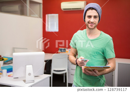 Young adult male standing and smiling in modern office, holding smartphone and folder with papers Young adult male standing and smiling in modern office, holding smartphone and folder with papers 128739117