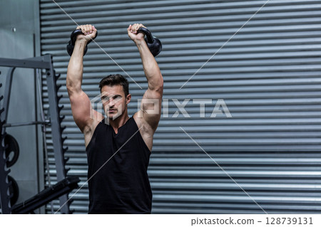 Man lifting kettlebells overhead at industrial-style gym, with weight rack and rubber flooring 128739131