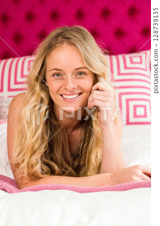 Woman lying on stomach in bedroom, smiling at camera with pink headboard and decorative pillows 128739135
