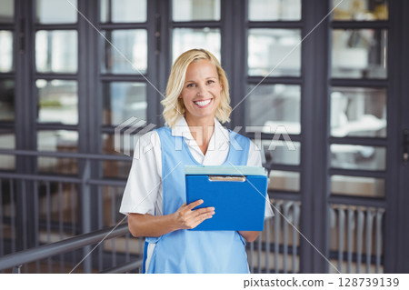young adult woman standing in office hallway holding bright blue clipboard with both hands, smiling 128739139