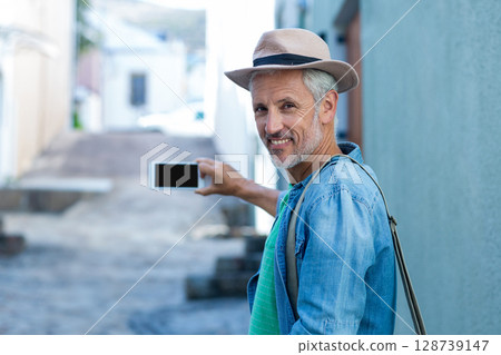 Senior man standing in sunlit stone-paved alley, holding smartphone and wearing fedora, copy space Senior man standing in sunlit stone-paved alley, holding smartphone and wearing fedora, copy space 128739147