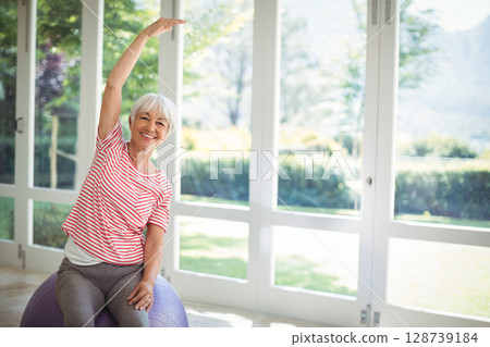 Senior woman performing side stretch on purple exercise ball in gym with mountain view, copy space Senior woman performing side stretch on purple exercise ball in gym with mountain view, copy space 128739184