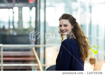Businesswoman turning toward camera in office with glass walls, desk plant and laptop, copy space 128739196