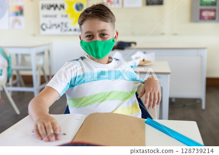 Schoolboy wearing green cloth mask writing in notebook in classroom, using pencil and blue ruler 128739214