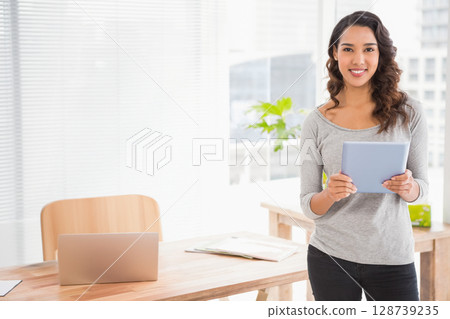 Woman standing in modern office holding tablet at wooden desk with laptop and documents, copy space 128739235