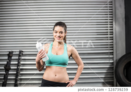 Woman standing and smiling at gym, holding white shaker bottle with weight rack, large tractor tire Woman standing and smiling at gym, holding white shaker bottle with weight rack, large tractor tire 128739239
