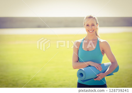 Mid-adult woman standing in sunny grassy field holding blue yoga mat, enjoying outdoor fitness Mid-adult woman standing in sunny grassy field holding blue yoga mat, enjoying outdoor fitness 128739241