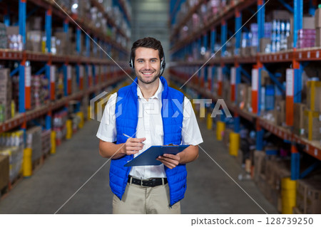Young adult man checking inventory in warehouse aisle with clipboard pen and headset, smiling 128739250