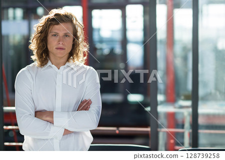 Young adult man standing with arms crossed in modern office space, with glass partitions, red beams Young adult man standing with arms crossed in modern office space, with glass partitions, red beams 128739258