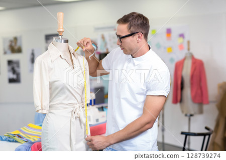 Male fashion designer measuring cream dress on torso mannequin in studio, with fabric rolls Male fashion designer measuring cream dress on torso mannequin in studio, with fabric rolls 128739274