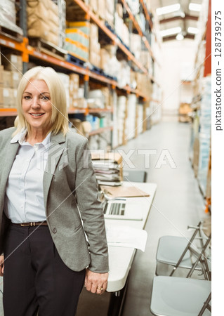 middle-aged woman standing beside work table in warehouse, checking boxes and paperwork with laptop 128739275