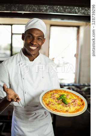African American male chef smiling and presenting freshly baked pizza in commercial kitchen African American male chef smiling and presenting freshly baked pizza in commercial kitchen 128739296