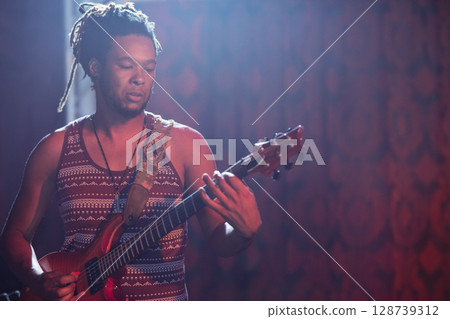Male musician playing electric guitar on dim stage under colored lights, with decorated strap Male musician playing electric guitar on dim stage under colored lights, with decorated strap 128739312