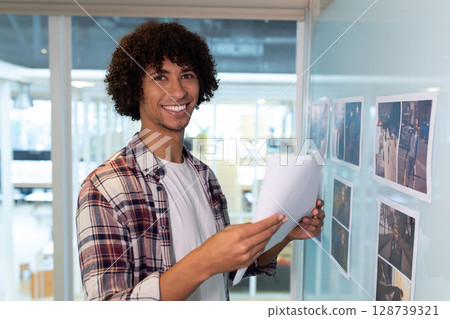 Man holding papers standing beside glass board reviewing printed photographs in office, copy space Man holding papers standing beside glass board reviewing printed photographs in office, copy space 128739321