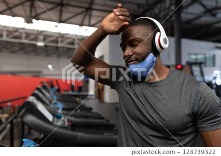 African American man wiping sweat on treadmill at gym with blue towel and mask, copy space 128739322