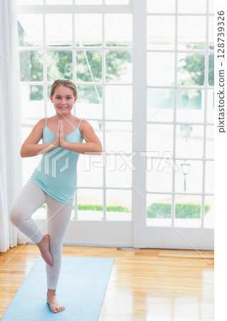 Child girl performing tree pose in living room with French doors, with light blue yoga mat Child girl performing tree pose in living room with French doors, with light blue yoga mat 128739326