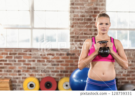 Fit woman lifting kettlebell in gym studio with stacked weight plates and exercise ball, copy space 128739331