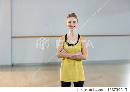 Young woman smiling and standing with crossed arms in dance studio, with ballet barre 128739345