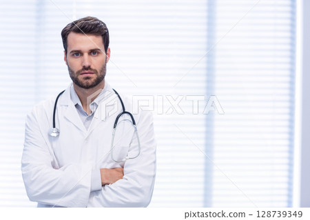 Male doctor standing with arms crossed in clinic room wearing lab coat and stethoscope, copy space 128739349