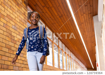 Smiling African American girl walking down brightly lit school hallway, with backpack Smiling African American girl walking down brightly lit school hallway, with backpack 128739364