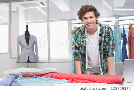 Smiling male fashion designer leaning on worktable in studio, with measuring tape and fabric rolls Smiling male fashion designer leaning on worktable in studio, with measuring tape and fabric rolls 128739373