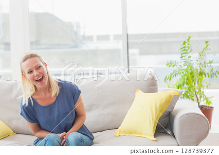 Mid-adult woman laughing, leaning on sofa by windows with yellow pillows in living room, copy space Mid-adult woman laughing, leaning on sofa by windows with yellow pillows in living room, copy space 128739377