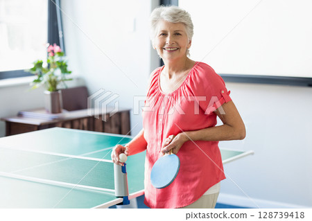 Smiling senior woman holding paddle and ball in game room, with ping-pong table and large window Smiling senior woman holding paddle and ball in game room, with ping-pong table and large window 128739418