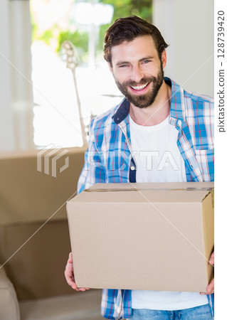 Smiling man holding cardboard box in living room, with acoustic guitar and couch Smiling man holding cardboard box in living room, with acoustic guitar and couch 128739420