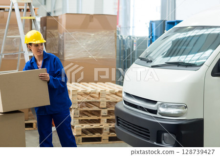 Lifting female worker carrying cardboard box in loading area, with delivery van, copy space 128739427