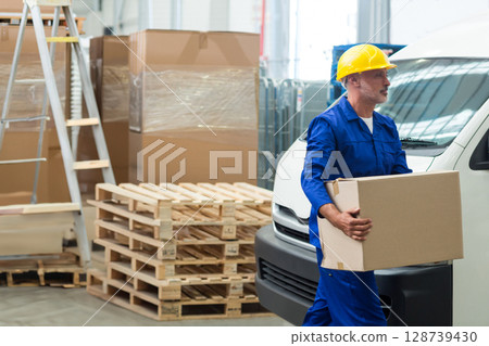 Walking woman carrying sealed cardboard box past pallets in warehouse loading area, copy space Walking woman carrying sealed cardboard box past pallets in warehouse loading area, copy space 128739430