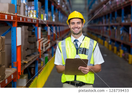 Inspecting male worker navigating wide warehouse aisle, with clipboard, safety vest and hard hat 128739433