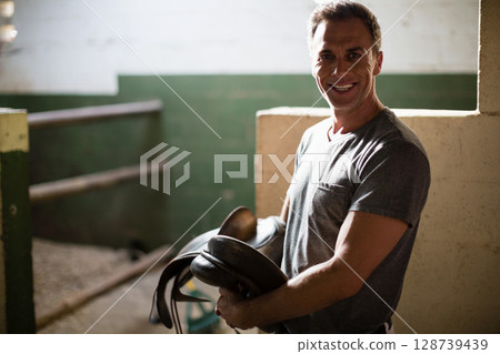 Smiling mature man holding dark leather saddle in barn aisle, with stable background, copy space 128739439