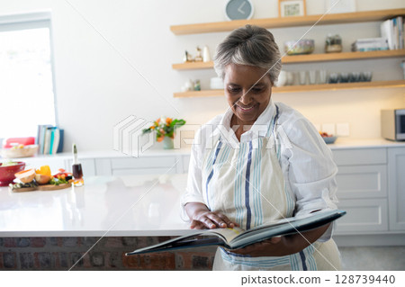 Senior African American woman holding open cookbook at kitchen island, with flowers and produce 128739440