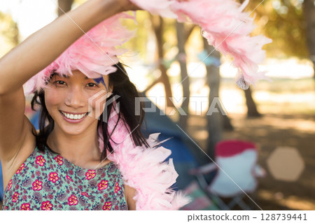 Holding pink feather boa above head, Asian woman dancing in wooded park, with tent and chair Holding pink feather boa above head, Asian woman dancing in wooded park, with tent and chair 128739441