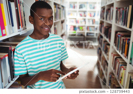 African American teenage boy tapping white tablet computer in library aisle, with tall bookshelves African American teenage boy tapping white tablet computer in library aisle, with tall bookshelves 128739443