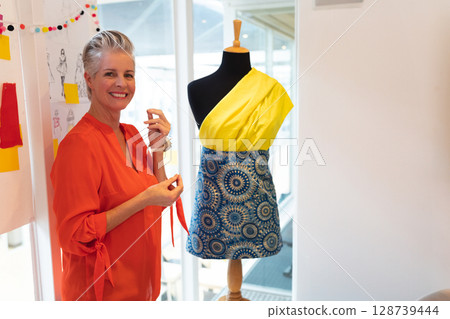 Smiling senior woman draping red fabric on dress form in design studio, with sketches, copy space 128739444
